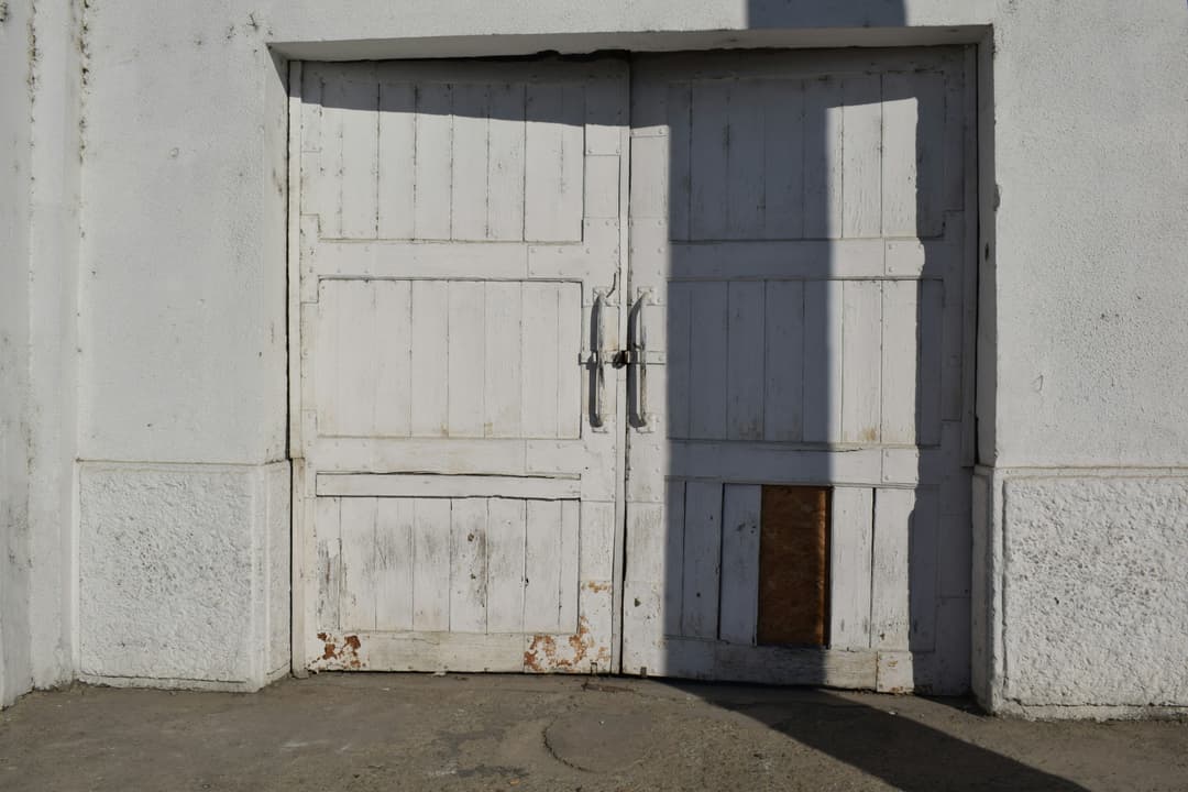 Weathered white wooden doors with rust and a patch, set against a textured wall.