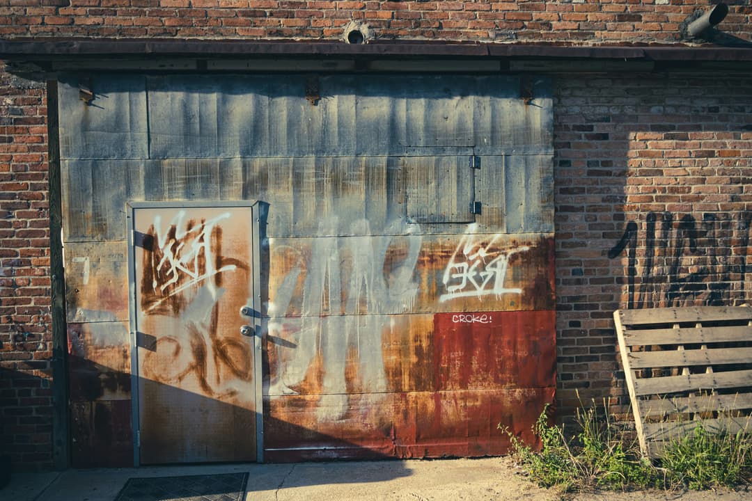 Rusty metal door with graffiti on a weathered brick wall and wooden pallet nearby.