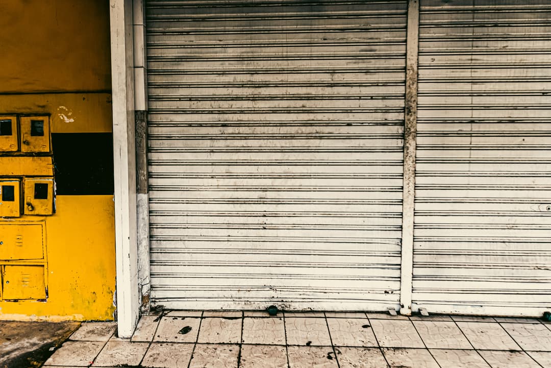 Closed metal shutter at a storefront with yellow wall and tiled sidewalk.