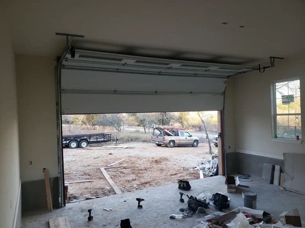 Garage interior with open door, revealing a vehicle and trailer outside. Construction materials visible.