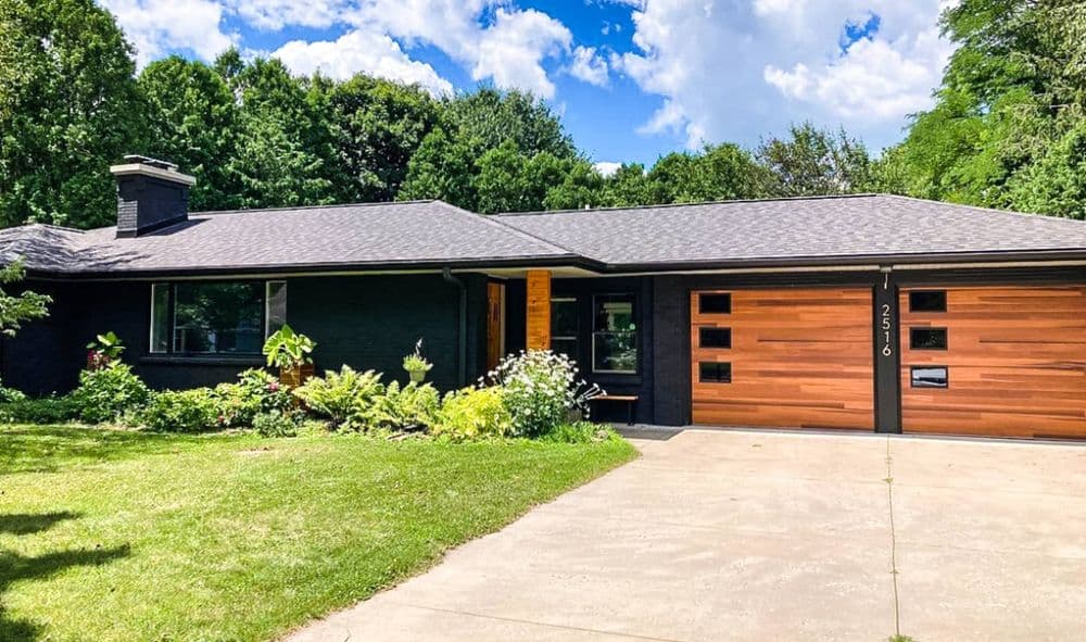 Modern black brick house with wood garage doors and lush landscaping under a blue sky.
